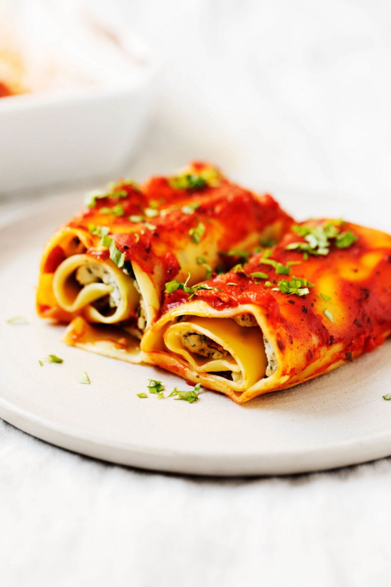 Two long, flat noodles have been filled with plant-based ricotta and spinach, topped with sauce, and rolled up.