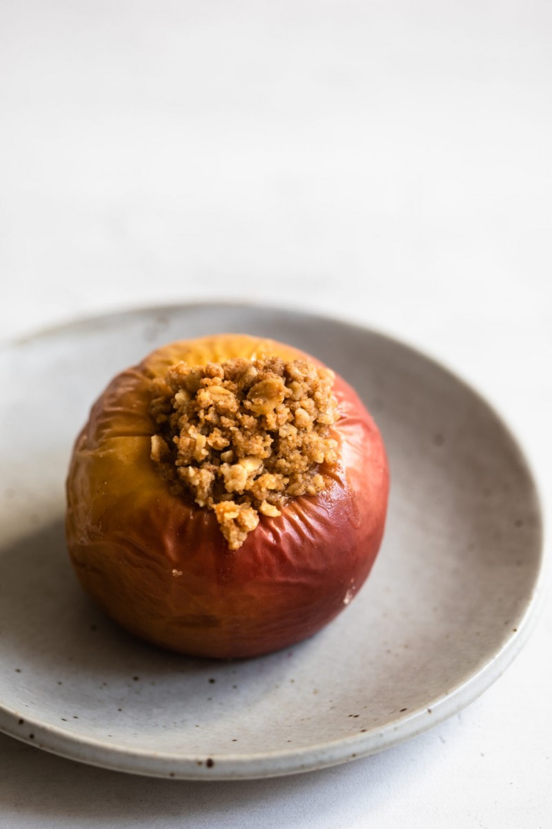 A single baked, stuffed red and golden apple is resting on a light gray, ceramic dessert plate.