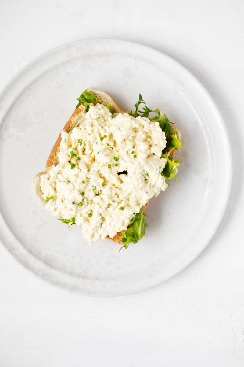 A slice of toast has been covered in a vegan tofu egg salad and a single green slice of lettuce. It rests on a white ceramic plate.