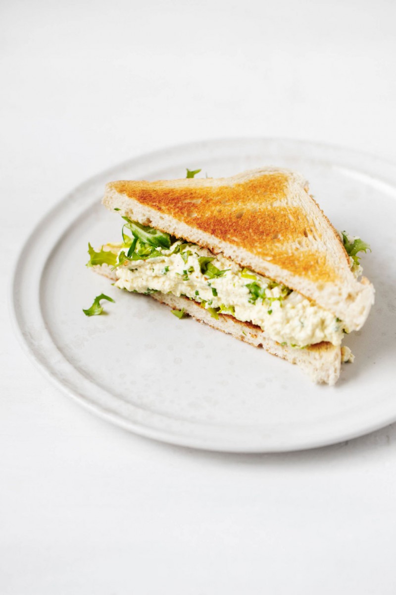 An angled photograph of a white plate that has been topped with half of a tofu egg salad sandwich and fresh greens.