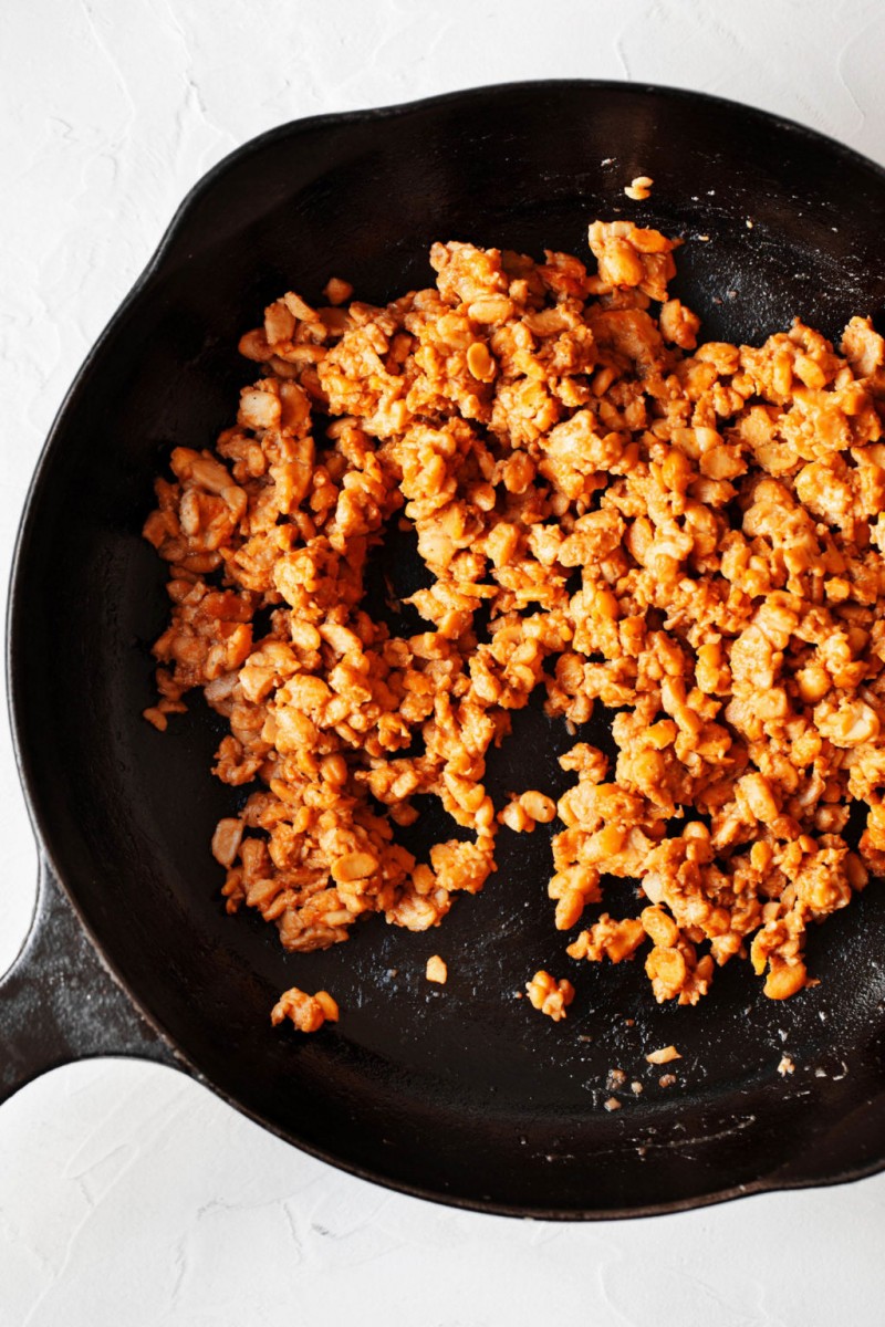 A black, cast iron skillet is filled with crumbled and seasoned tempeh.