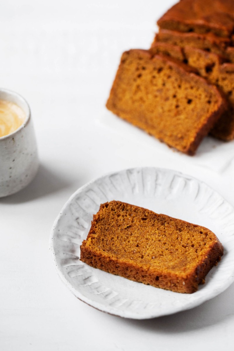 A piece of pumpkin bread, ready for snacking along with a frothy latte. And slices of more bread in the background.