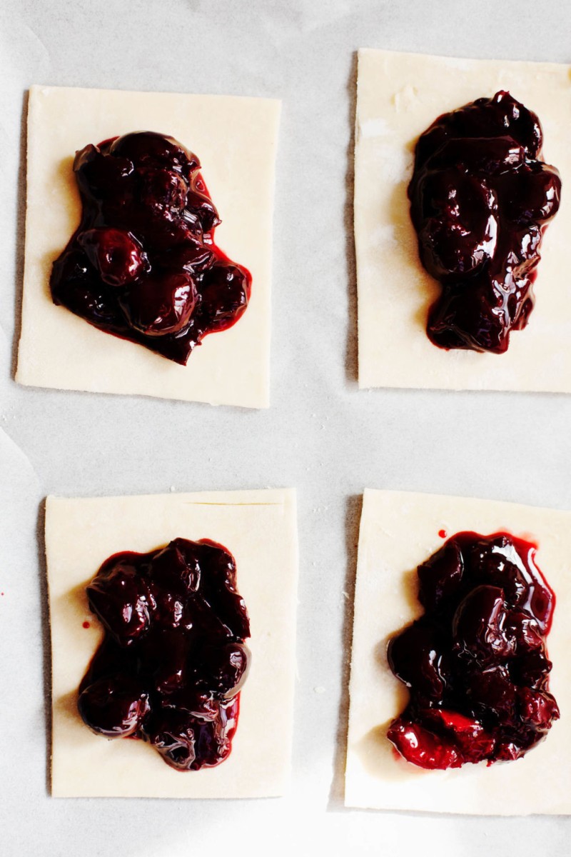 Vegan pastry pies on a parchment sheet, cut into rectangles. Each rectangle is topped with a sweet cherry filling.