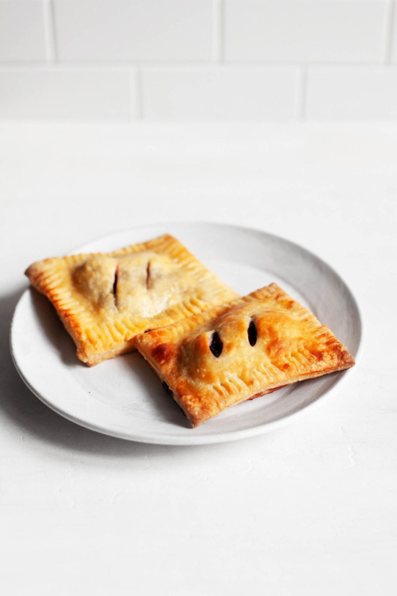 Two rectangular, vegan cherry hand pies with crispy golden pastry sit next to each other on a dessert serving plate.