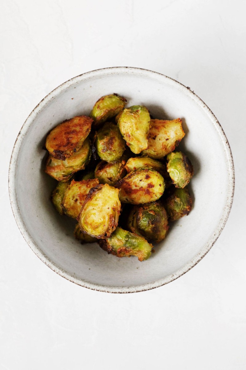 A white, small bowl holds a number of glazed, halved, and crispy roasted Brussels sprouts.