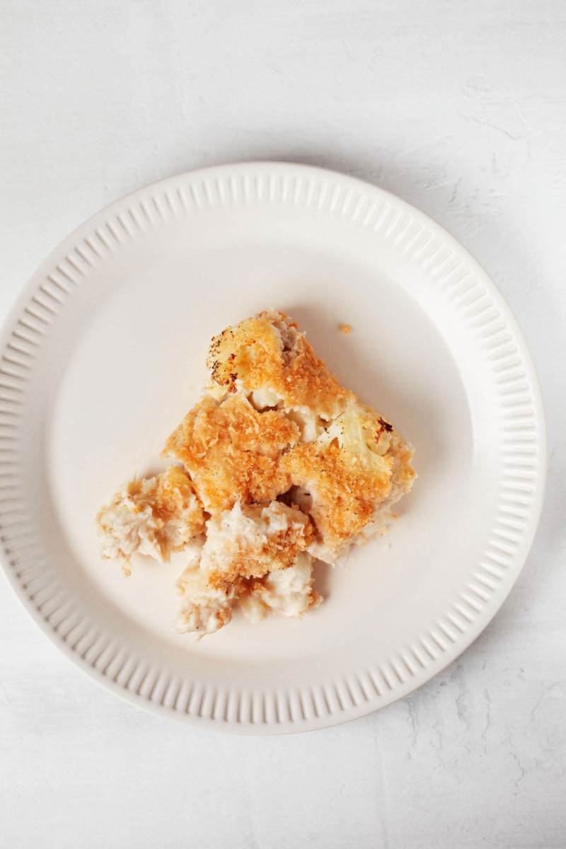 A single slice of a plant-based cauliflower gratin rests on a fluted white serving plate. It's topped with bread crumbs that have turned golden brown in the oven.