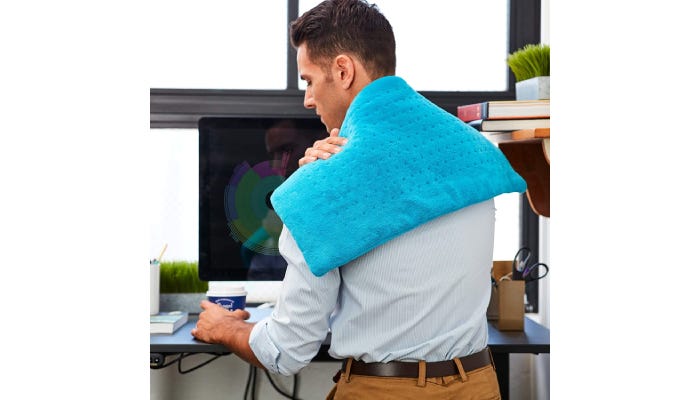 A man with his back to the camera, wears a light blue heating pad across his back shoulders while standing in an office. He holds the heating pad with his right hand across his left shoulder.