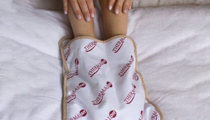 A woman's hands linger above the heating pad into which she's slipped her feet. The background is a white comforter and the heating pad has multiple Thermalon logos printed in red across it. 