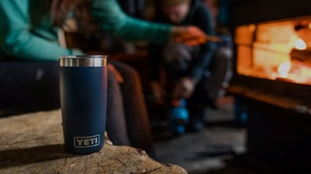 A Yeti 20 oz. tumbler sitting on a tree stump next to a group of people roasting food on a fire. 