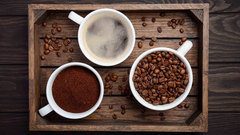 Whole coffee beans, ground coffee and a cup of hot brewed coffee placed on a rustic wooden tray with various beans spread on the tray.