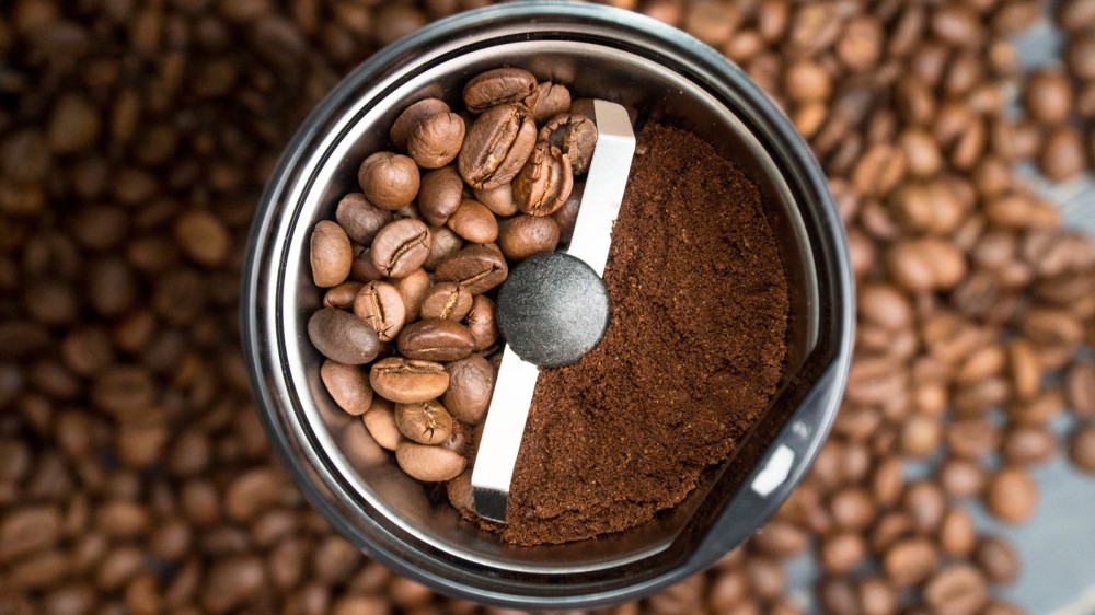 A blade coffee grinder half filled with beans, and the other half filled with ground coffee with a lot of coffee beans in the background.