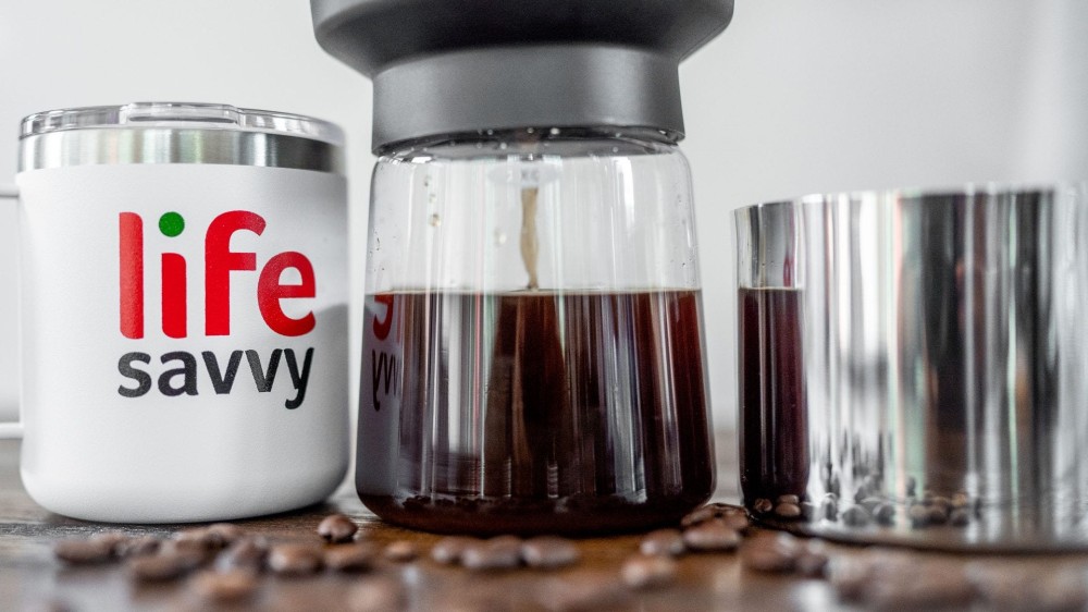 white coffee mug next to a cold brew coffee maker and metal coffee grounds cup