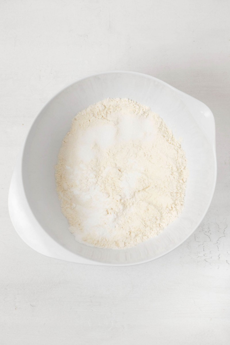 A white mixing bowl contains a mixture of flour and other dry ingredients. It rests on a white surface.