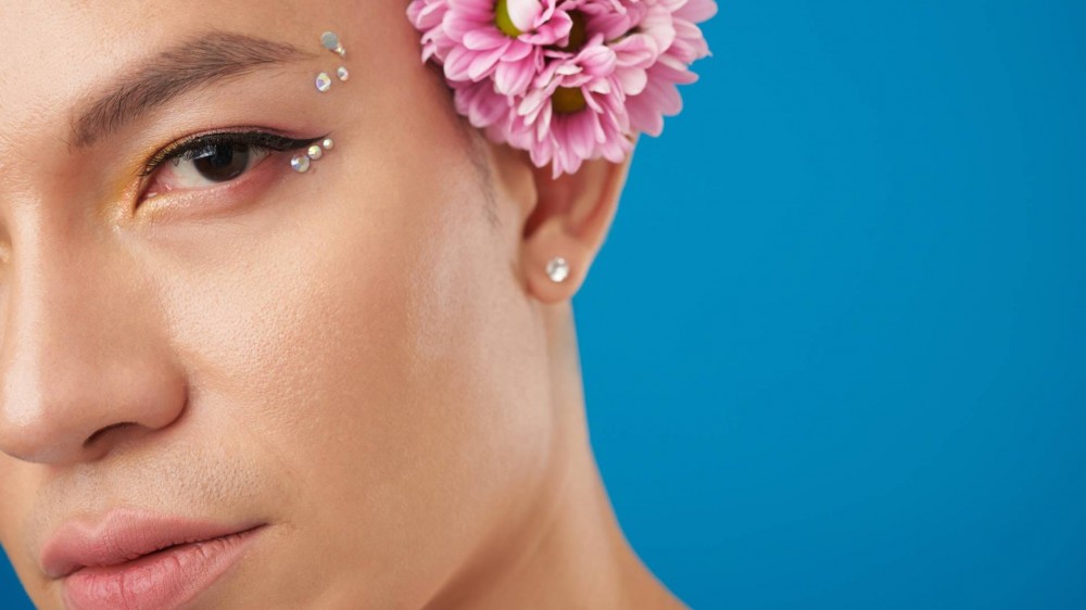 Face of serious young man with eye makeup, rhinestones on face, and flowers behind ear.