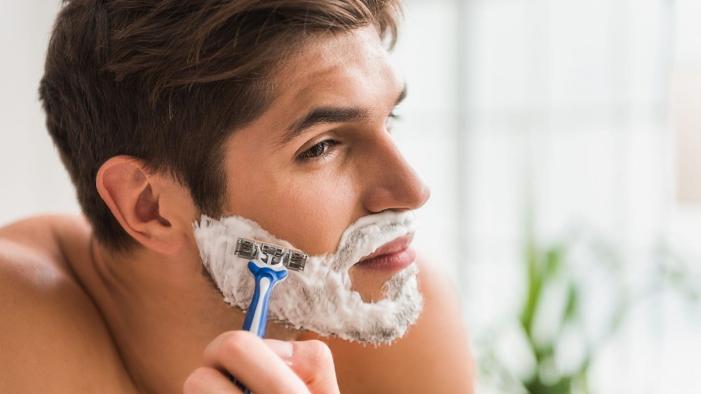 A man shaves his beard with a disposable razor and shaving cream.