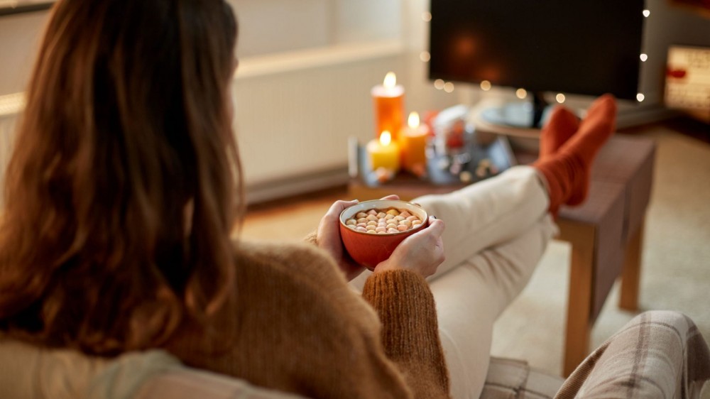 Young woman watching tv and drinking hot chocolate with marshmallow with her feet on table at cozy home.