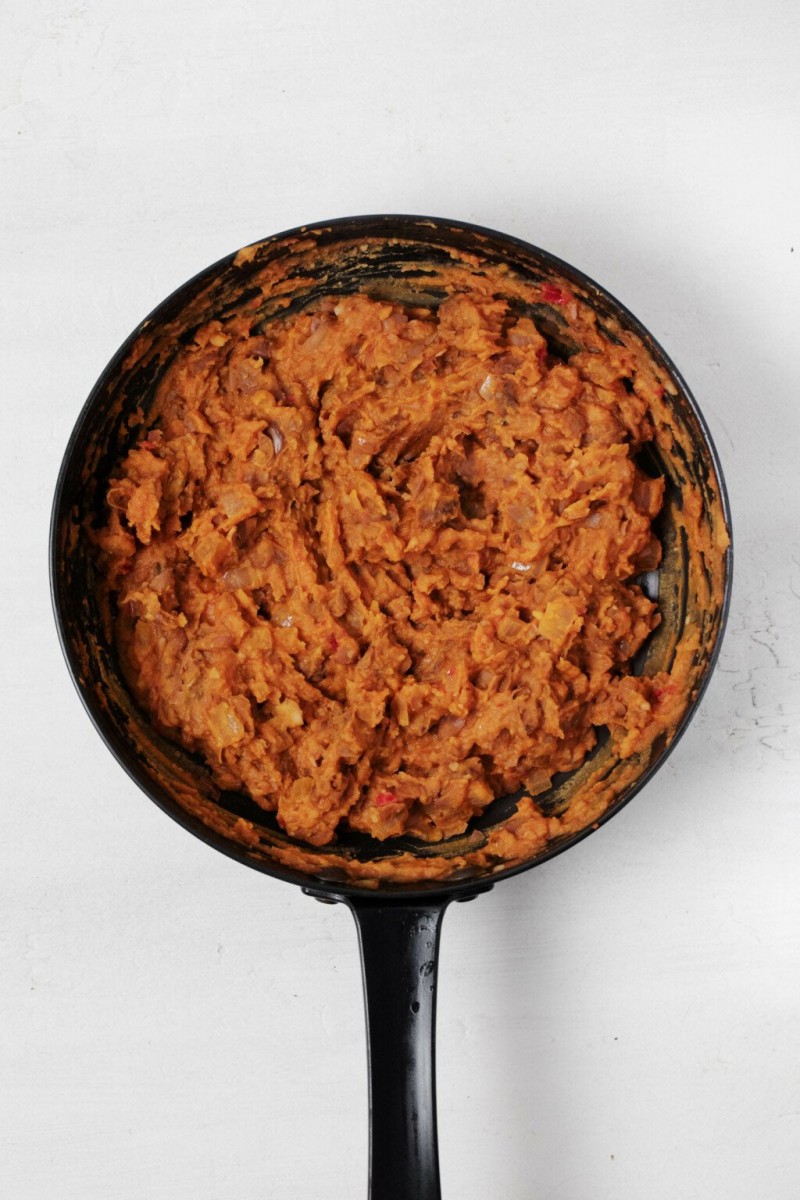 A round, black cast-iron skillet is being used to make vegan refried beans. It rests on a white surface.