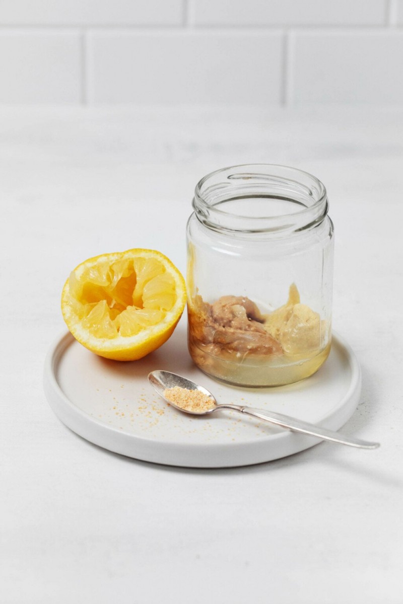 A jar with salad dressing ingredients, a half lemon, and a spoonful of mustard are resting on a round white plate.