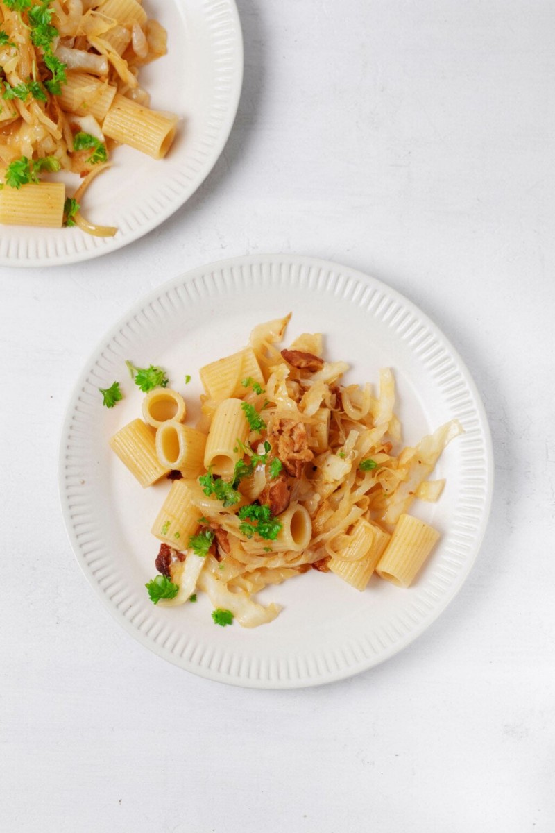 Two plates of a vegan cabbage and onion rigatoni dish are resting on a white surface. The pasta is garnished with flecks of green parsley.