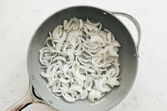 White onions are thinly sliced and about to be sautéed in a gray frying pan.