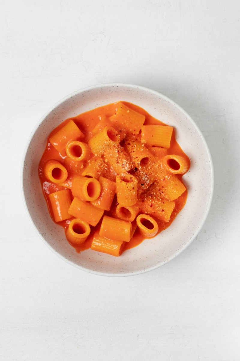 A creamy, vegan red pepper pasta is served in a round, white bowl. The bowl rests on a white surface.