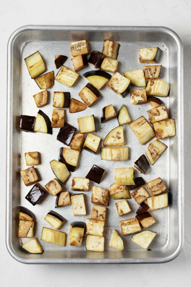 Roasted eggplant cubes are pictured on a rimmed, silver baking sheet.