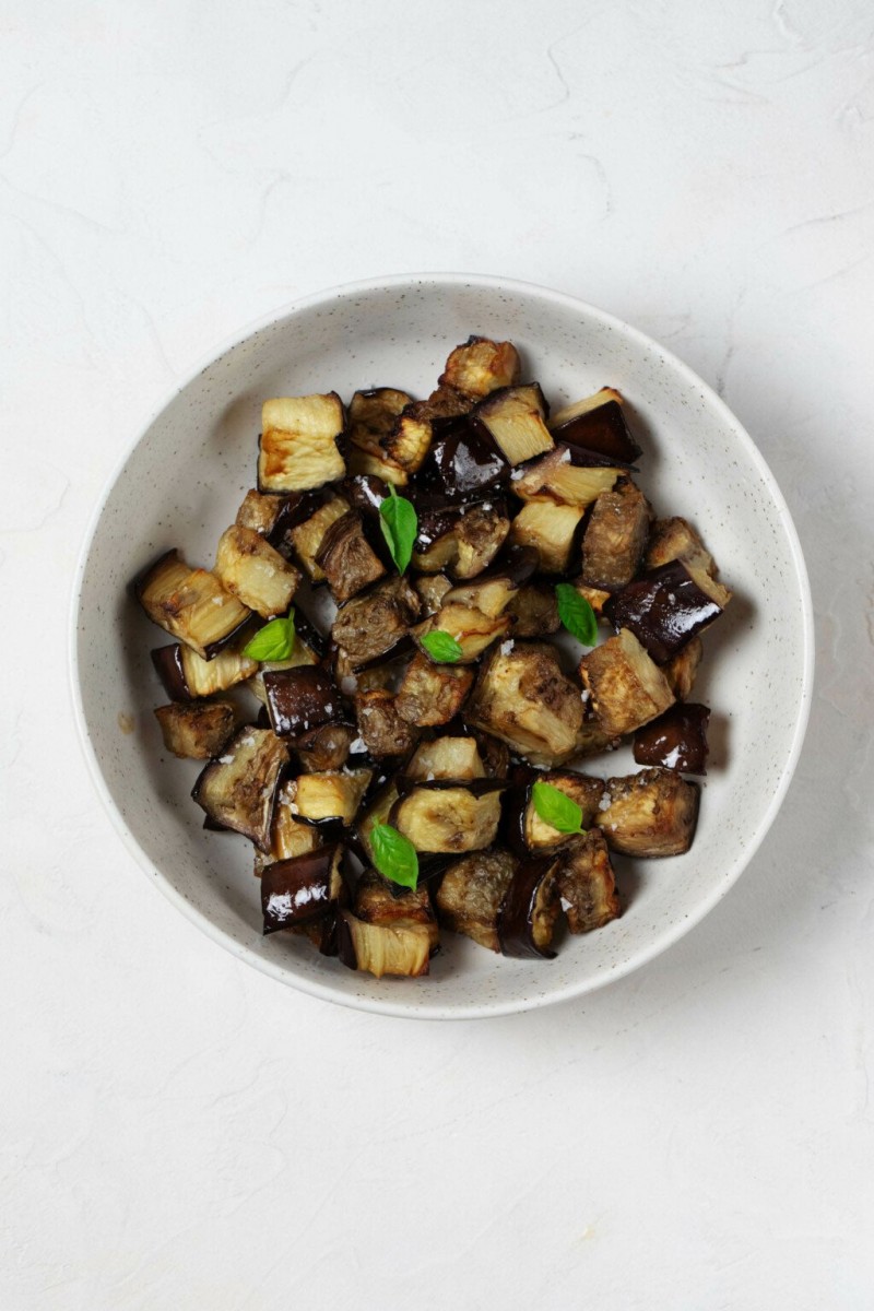 A white, ceramic bowl is filled with roasted vegetables and tiny leaves of fresh, green basil.