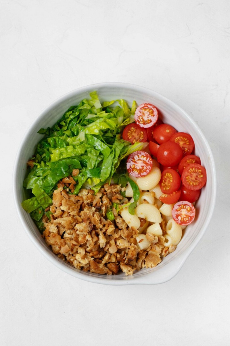 A large, round white mixing bowl is filled with tomatoes, tempeh bacon, cooked pasta, and chopped romaine lettuce.