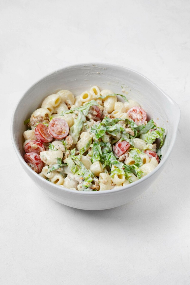 A white mixing bowl is filled with the ingredients for a vegan BLT pasta salad, including pasta, romaine lettuce, and tomatoes.