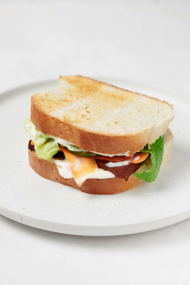 A sandwich, made with toasted white bread, is pictured on a round, white plate. The sandwich is topped with mayonnaise, pickled vegetables, and slices of baked tofu.