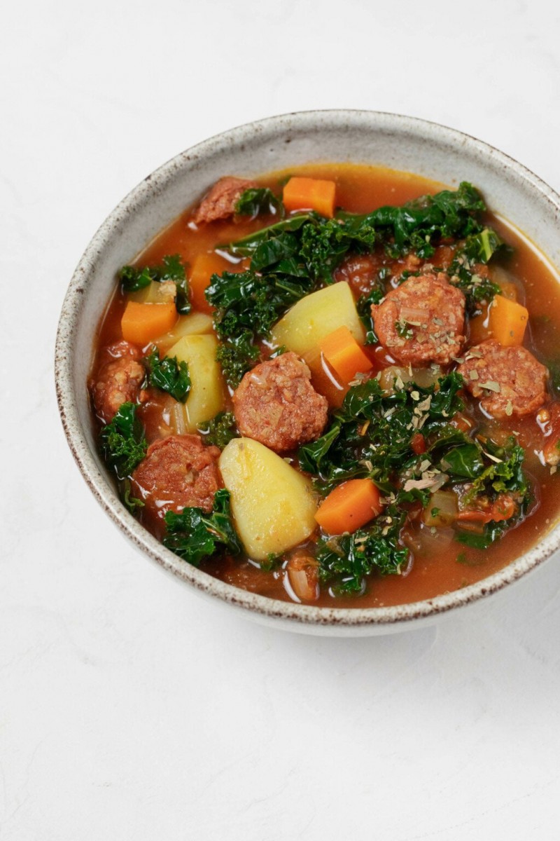 A gray ceramic round bowl has been filled with a hearty plant-based soup. The soup is chock full of kale, potatoes, and vegan sausage slices.