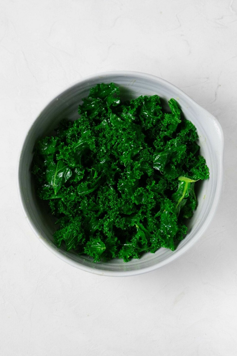 An overhead image of a white mixing bowl. The bowl is filled with raw kale, which has been marinated with a vinaigrette.