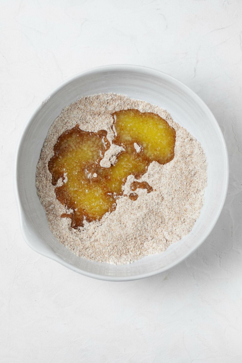 A white mixing bowl is filled with flour, cinnamon, and sugar. There's a small pool of melted butter visible in the center. 