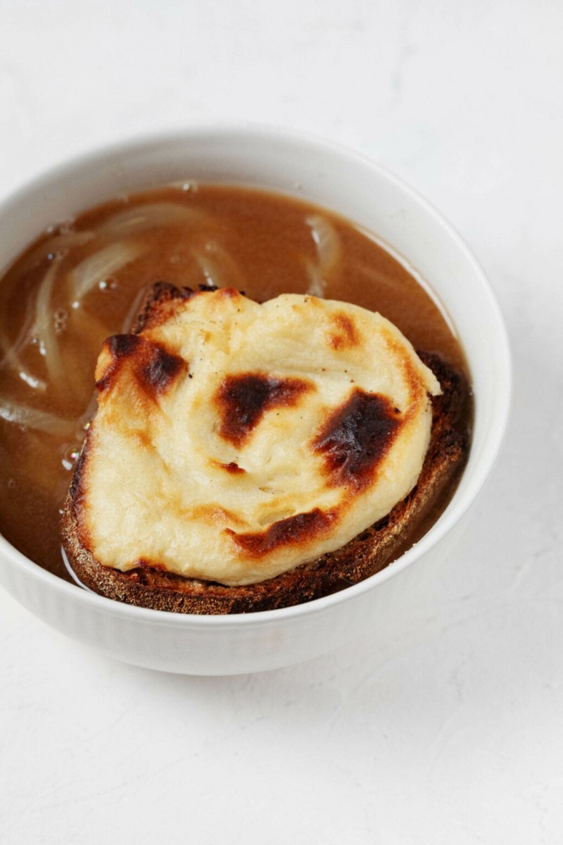 An overhead image of a round, white bowl of vegan French onion soup, which has been topped with a browned cheese toast. The toast rests directly in the bowl.