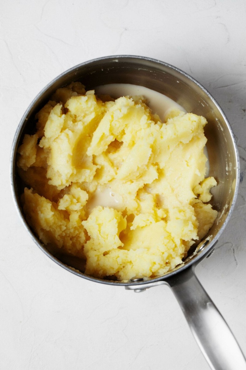 A silver saucepan is pictured from above. It contains mashed potatoes and warm milk.