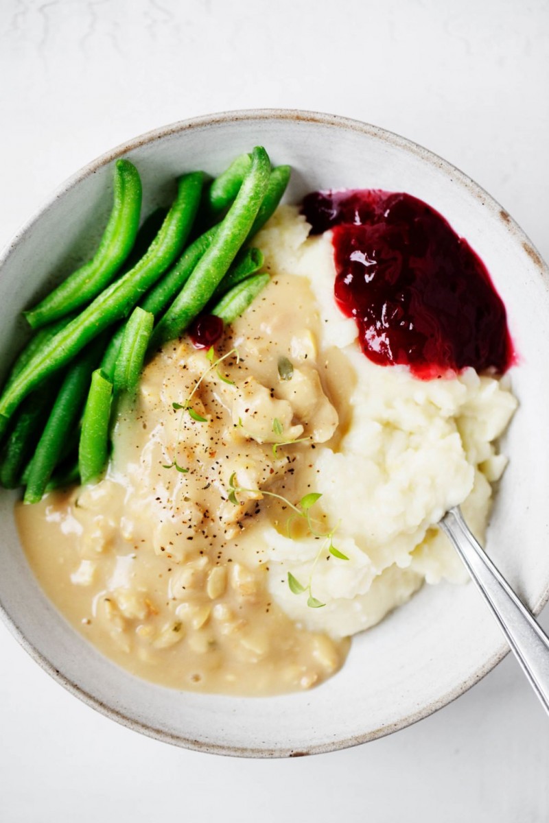 A bowl of vegan holiday food, including mashed potatoes, tempeh and gravy, green beans, and a little scoop of cranberry sauce.