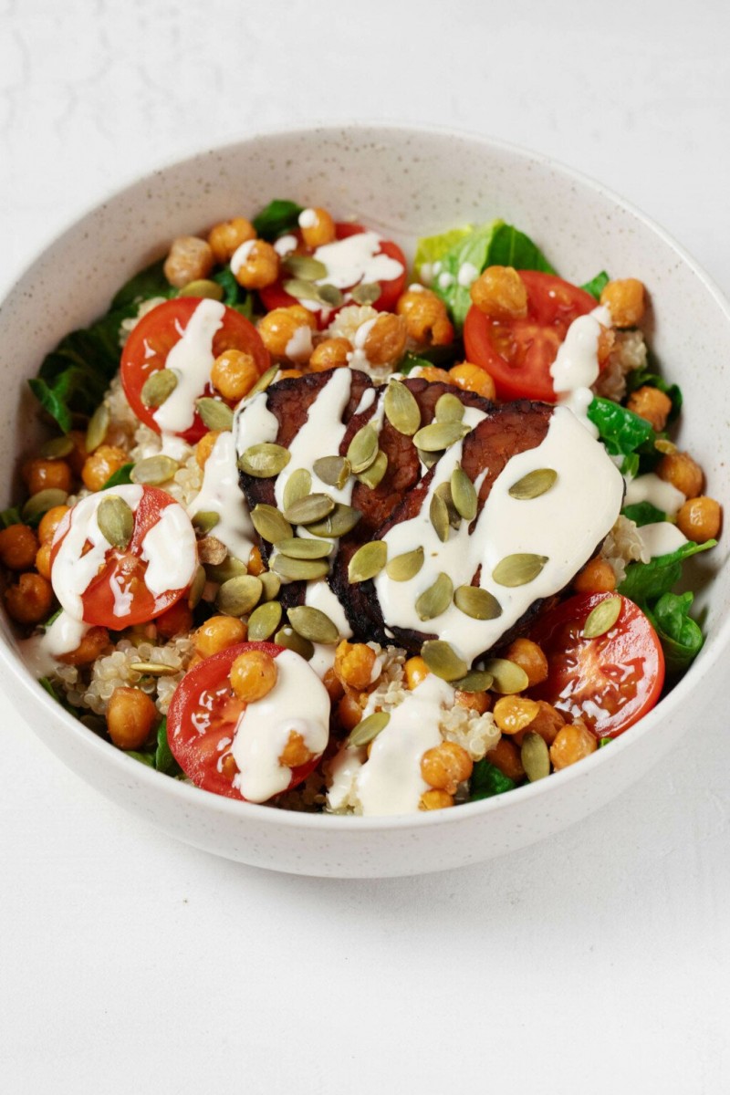 A large, white bowl is filled with vegetables, roasted chickpeas, quinoa, and a creamy ranch dressing. The bowl is topped with pepitas. 