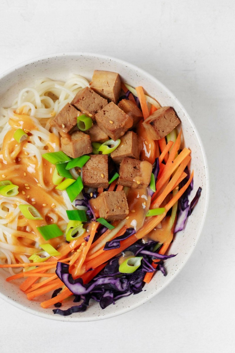 An overhead, close-up image of a white ceramic bowl. It's filled with julienned carrot and cabbage, tofu cubes, noodles, and a sauce.