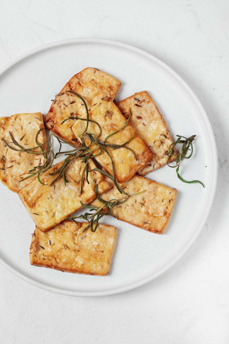 A round, white plate has been topped with golden baked tofu slices and rosemary leaves.