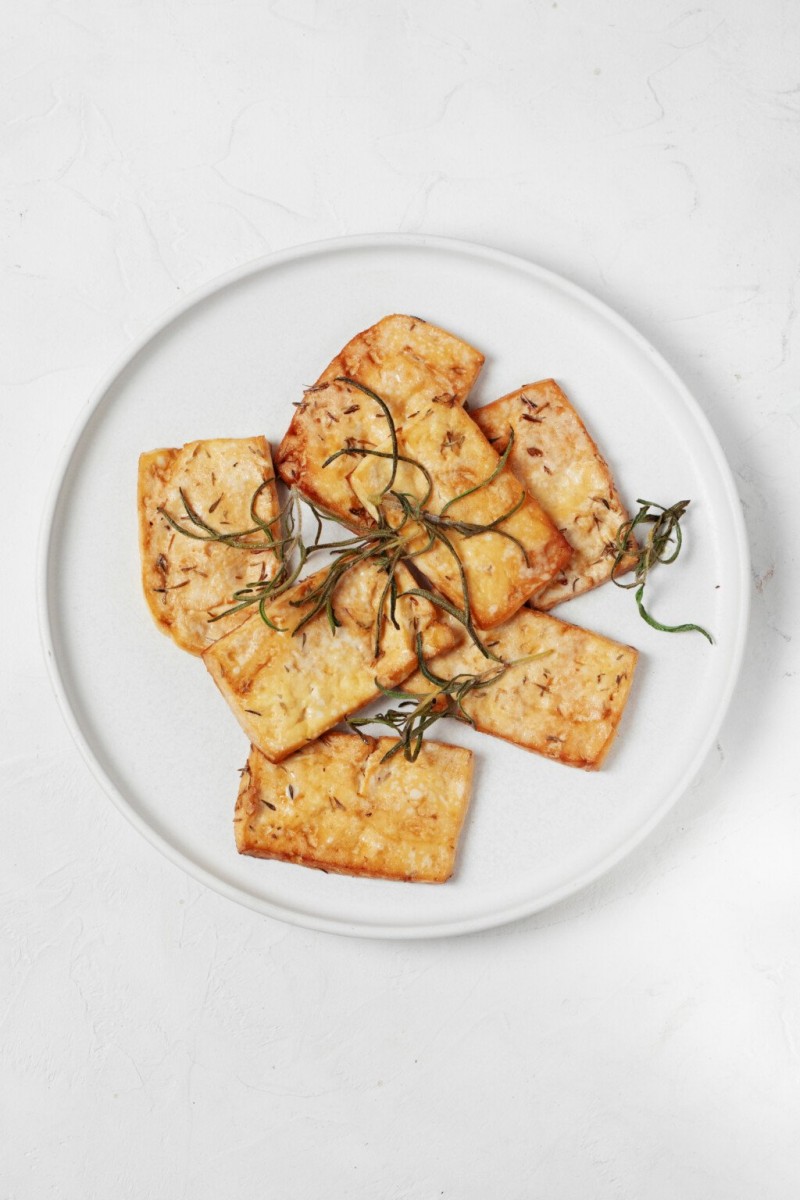 A round, white plate has been topped with golden baked tofu slices and rosemary leaves.