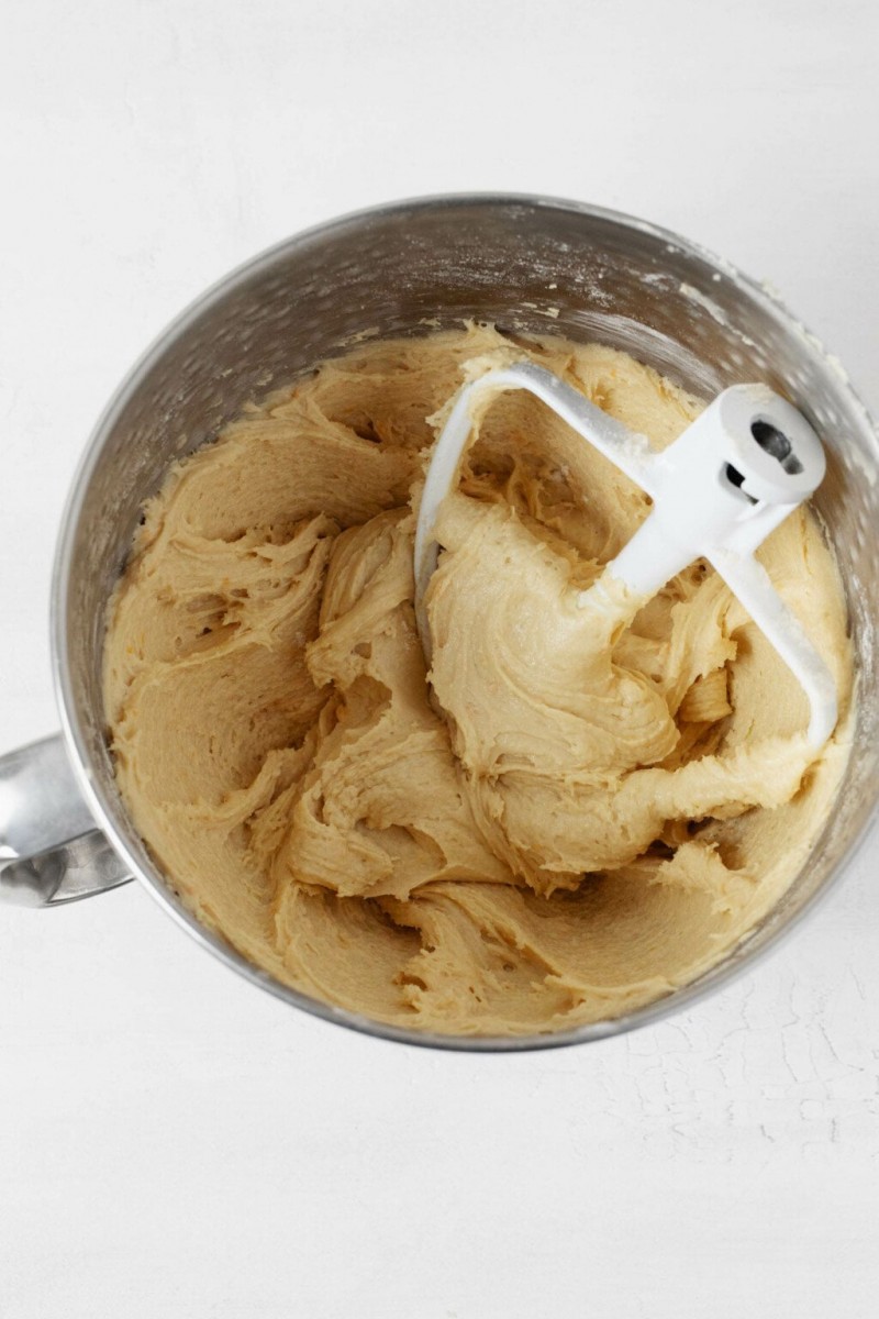 Pale yellow, creamed butter and sugar are pictured from above in a silver mixing bowl with a paddle attachment.