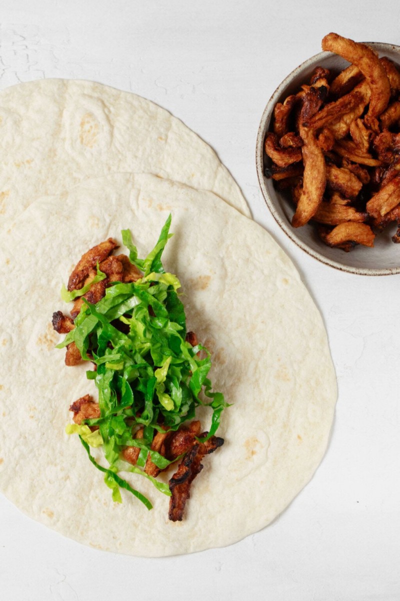 A vegan buffalo “chicken” wrap is mid-assembly on a work surface.