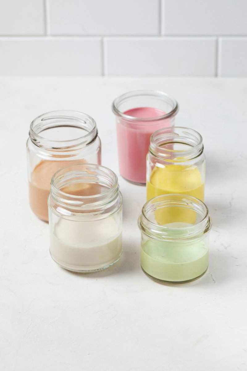 Five glass mason jars rest on a white surface. Each is filled with a different color of creamy tahini dressing.